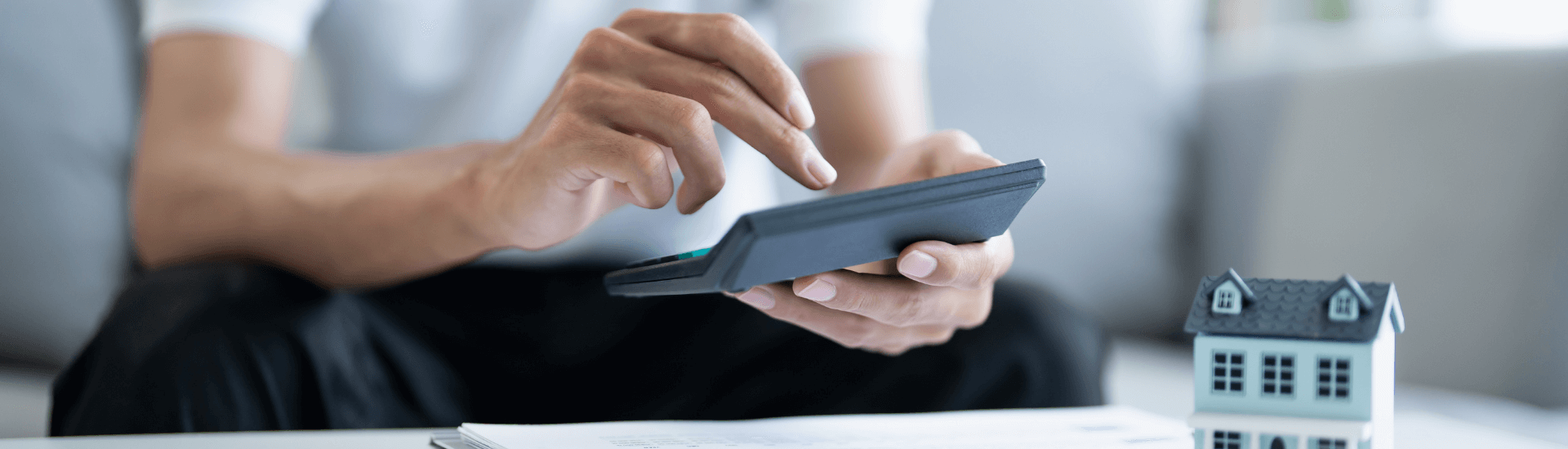 Mano che utilizza una calcolatrice vicino alla miniatura di una casa sul tavolo, concetto di cedolare secca per l’affitto